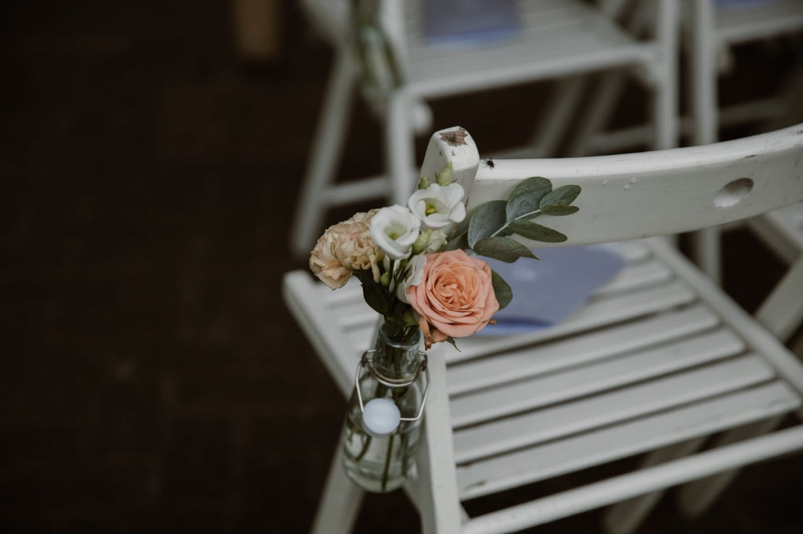 Corsage vrouw met pastelkleurige bloemen en subtiele groenaccenten, bloemen in vaas hangend aan witte klapstoel