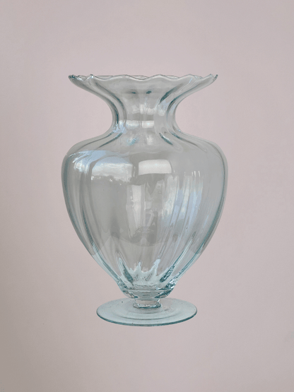 Grote glazen vaas met diepe ribbels, smalle bovenhals en ronde voet, met lichtroze achtergrond.