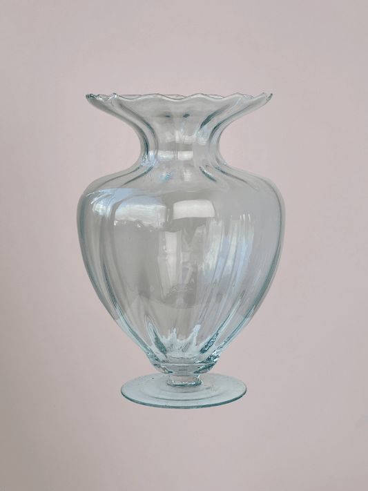 Grote glazen vaas met diepe ribbels, smalle bovenhals en ronde voet, met lichtroze achtergrond.